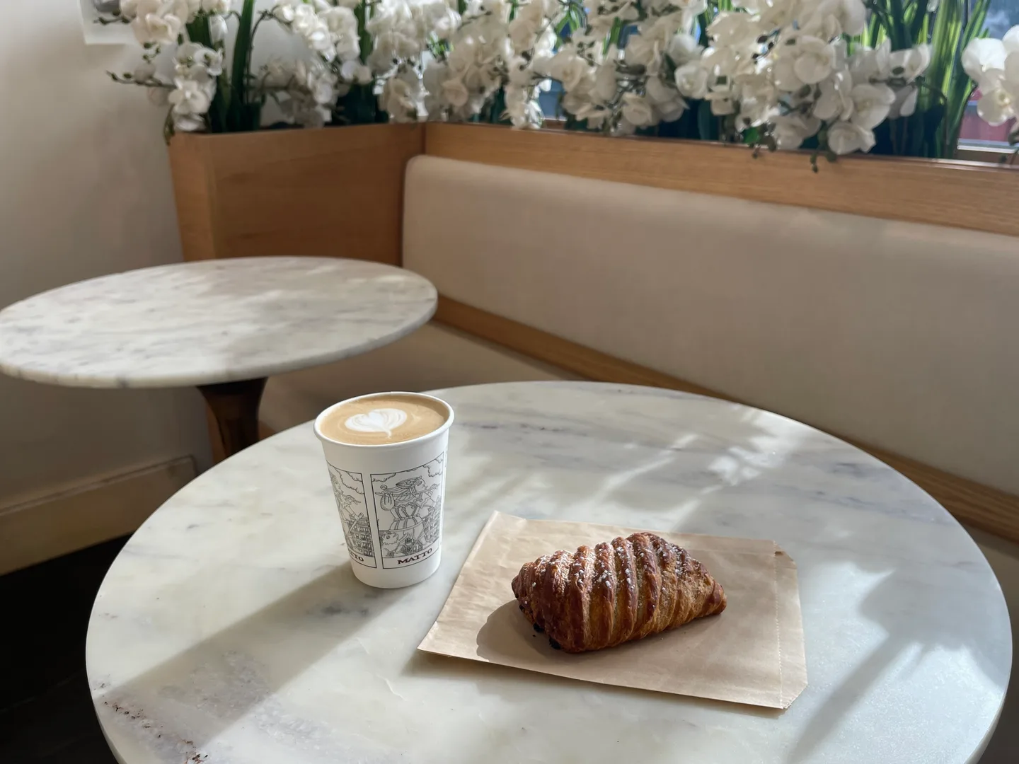 Coffee and a chocolate-drizzled pastry on a sunlit marble table beside café seating