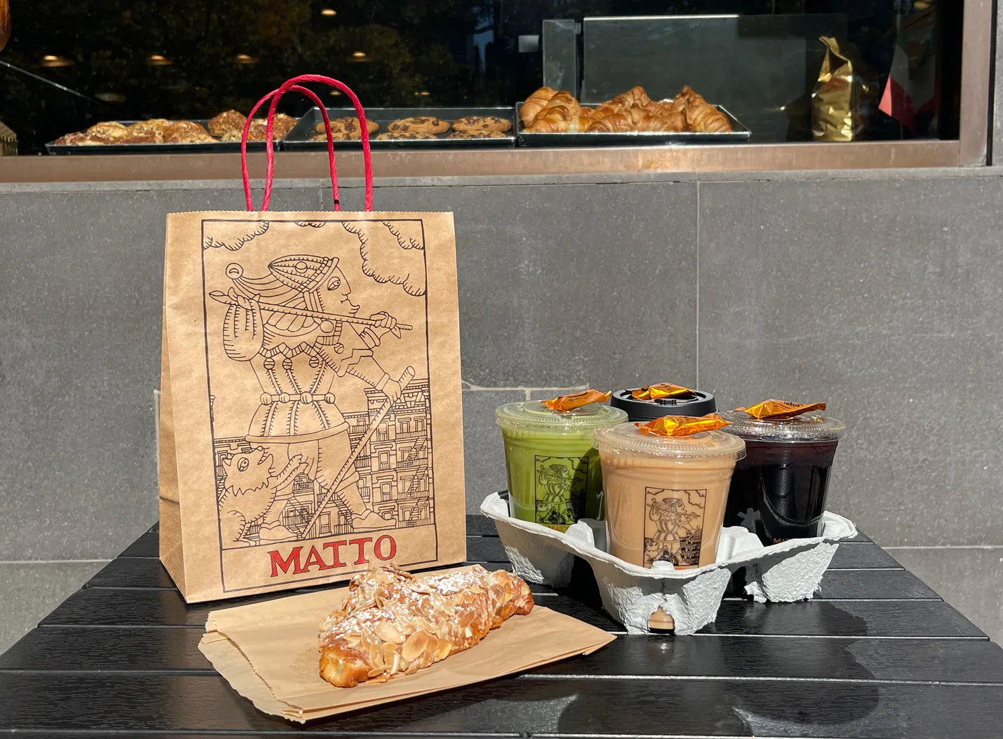 A Matto takeaway bag, iced drinks, and an almond croissant outside the café